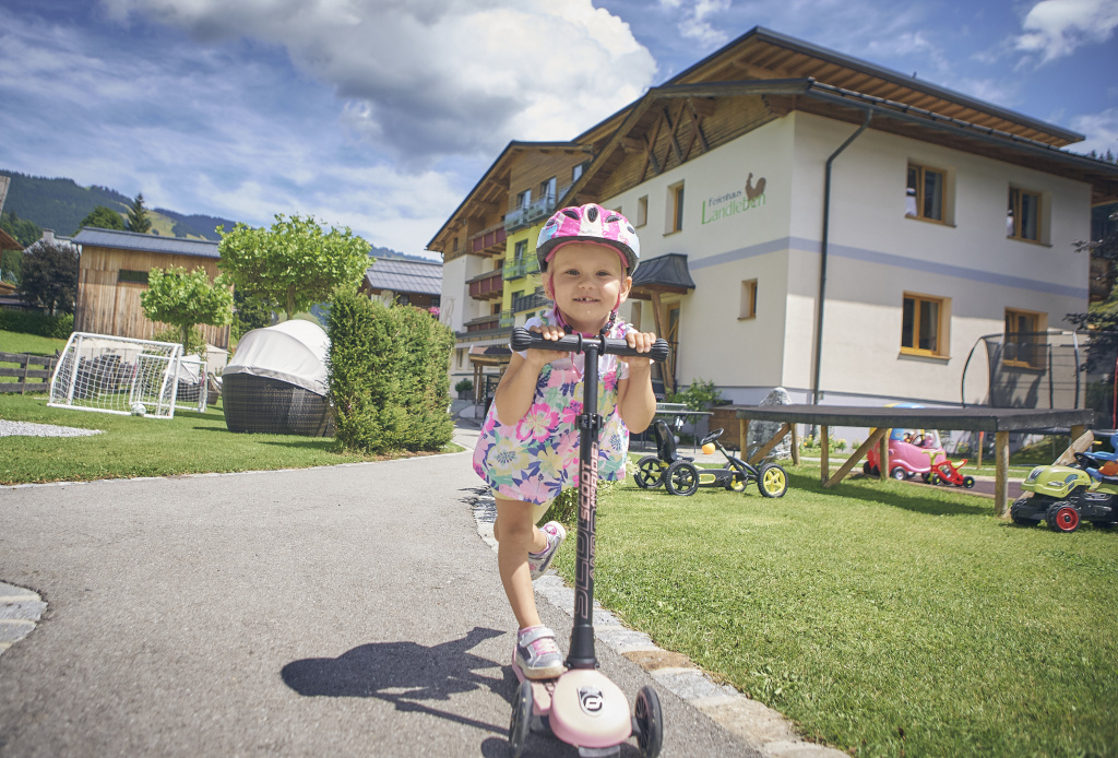 Kind mit rosa Helm fährt Roller vor einer Hotelanlage mit Spielbereich und Bergen im Hintergrund