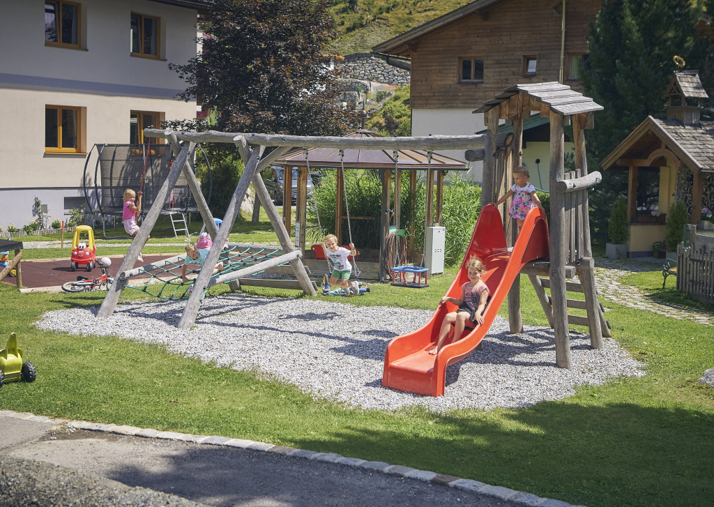 Kinder spielen auf einem Holzspielplatz mit Rutsche, Schaukel, Kletternetz und Trampolin im grünen Hotelgarten